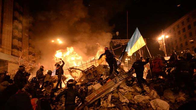 Picture of Ukraine riots in the streets with fire in the background and rioters climbing wreckage with one holding a Ukranian flag
