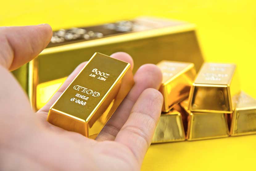 Hand holding 200 gram fine gold bar with stacks of larger gold bars in yellow background