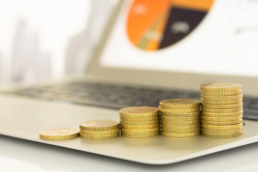Ascending stacks of gold coins sitting atop a laptop, with financial portfolio on computer screen in background