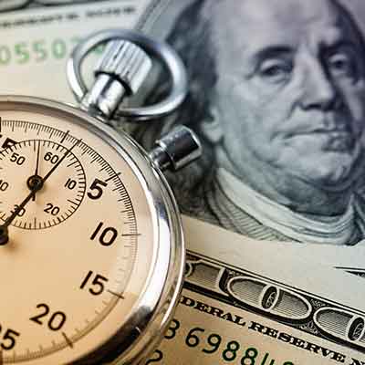 Silver pocket watch sitting on top of hundred dollar bills spread across a table.