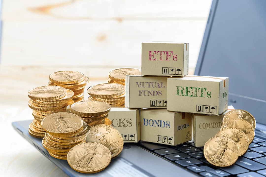 Stacks of gold coins and boxes with stocks, bonds, commodities, REITs, ETFs and mutual funds sitting on top of a laptop keyboard