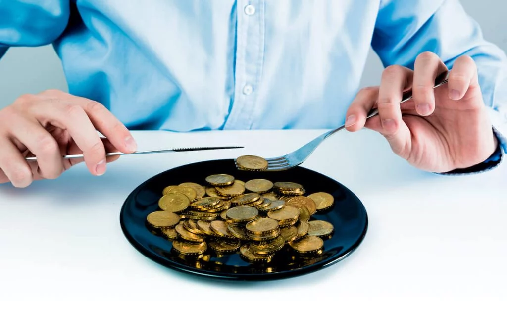 Man with knife and fork holding a cold coin from a plate with gold coins