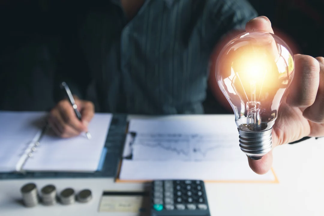 Person writing in notebook with graphs in front of him, coins, and a calculator holding an illuminated lightbulb