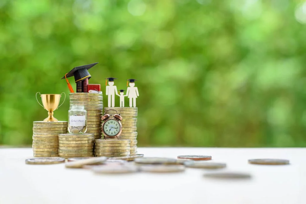 Stack of gold coins with coin jar labeled "Education", clock, trophy, graduation cap and books, and a family cutout with graduation caps on them
