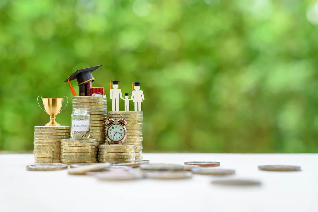 shutterstock_1447312808 Stack of gold coins with coin jar labeled "Education", clock, trophy, graduation cap and books, and a family cutout with graduation caps on them