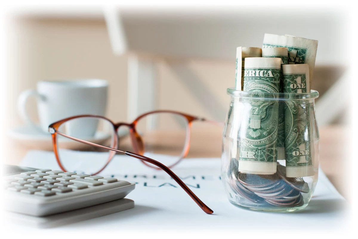 Glass jar containing cash next to glasses, calculator, and papers labeled “RETIREMENT PLAN”