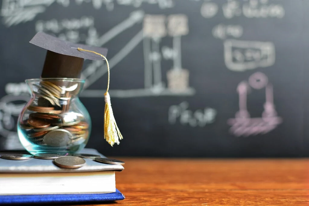 Jar filled with coins wearing graduation cap with educational chalkboard in background