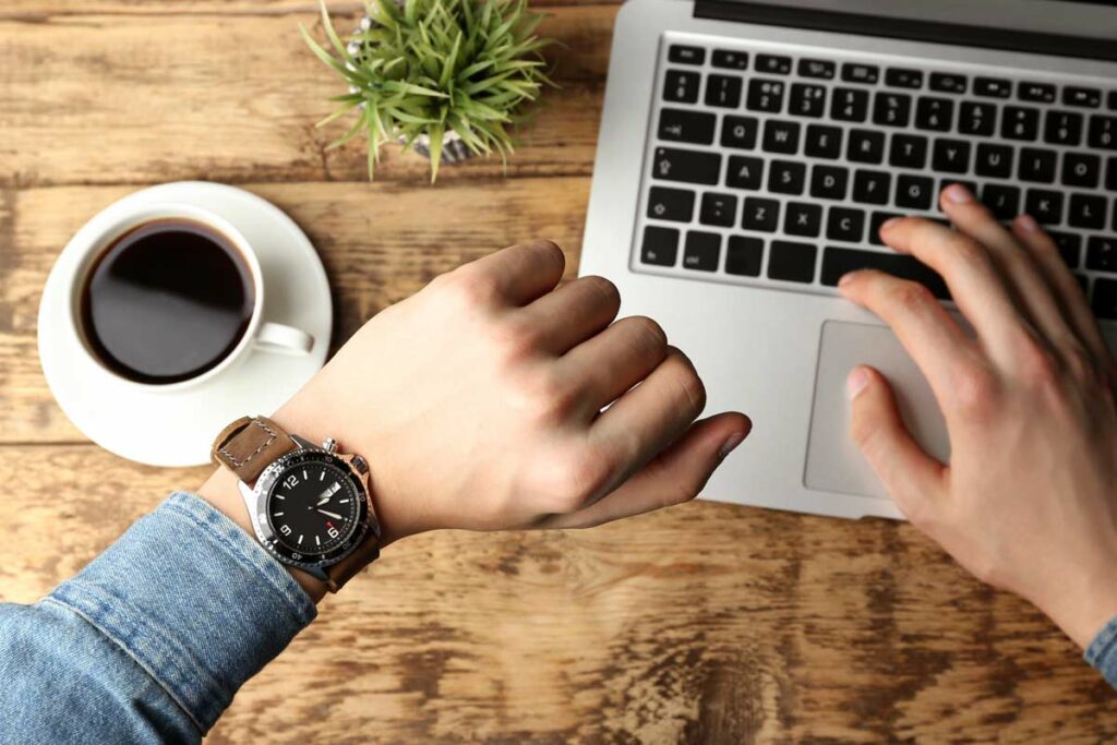 Looking at watch while working on laptop beside coffee