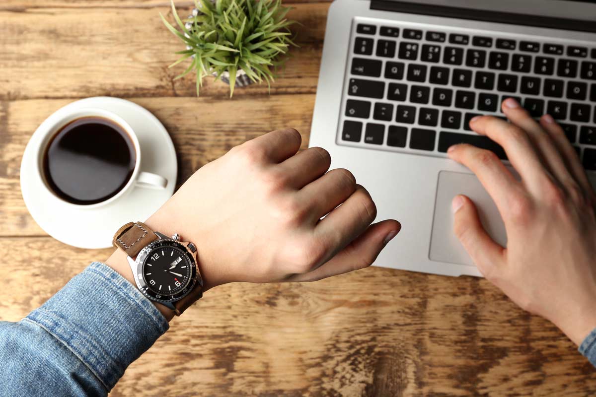 Looking at watch while working on laptop beside coffee