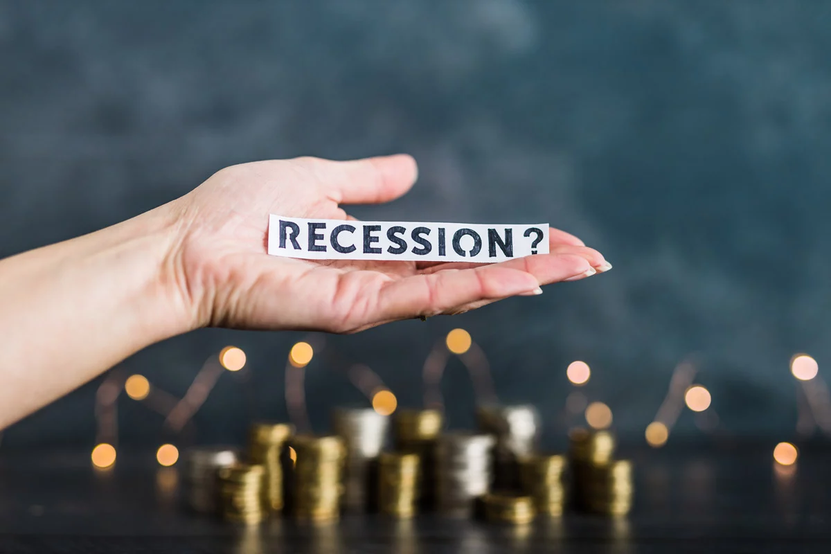 Hand holding paper reading "RECESSION?" with gold and silver coins in the background